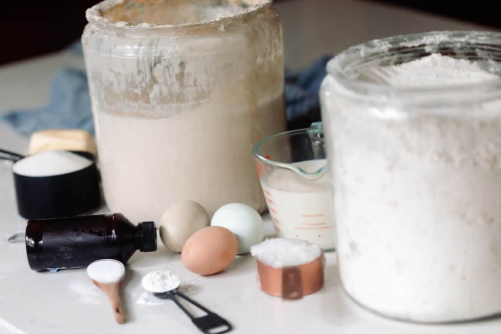 jars of flour and sourdough starter on a white countertop with eggs, and measuring cups of spoons of other ingredients