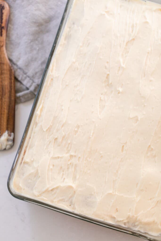 sourdough vanilla cake topped with fluffy buttercream frosting on a white quartz countertop with a wooden spoon and gray tea towel to the left