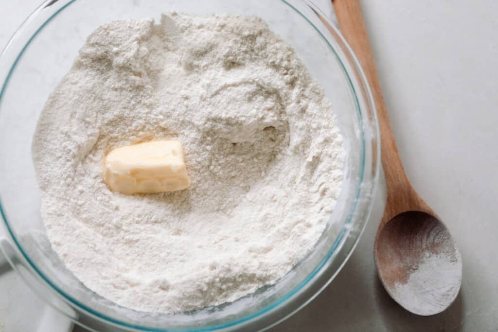 flour, sugar, baking soda, baking powder, and butter in a glass bowl with a wooden spoon to the right.