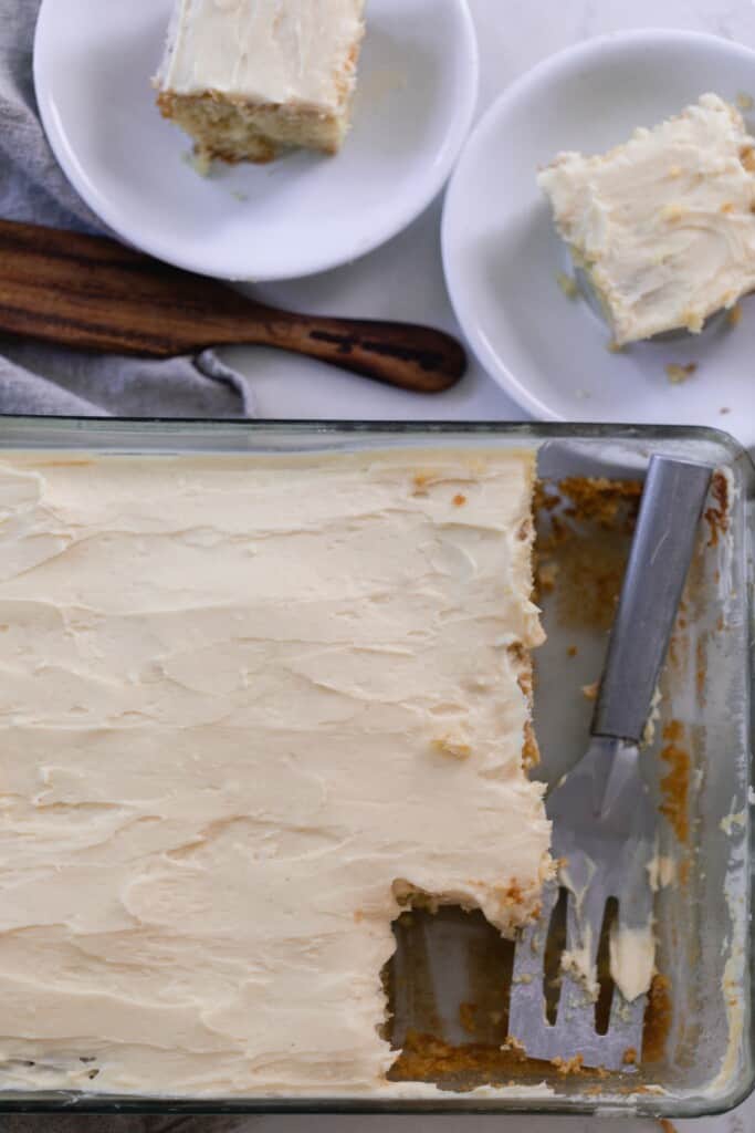 overhead photo of a baking dish with sourdough cake with a thick layer of buttercream frosting. A metal spatula is resting in the glass baking dish. Two square pieces of cake on white plates sit next to the baking dish.