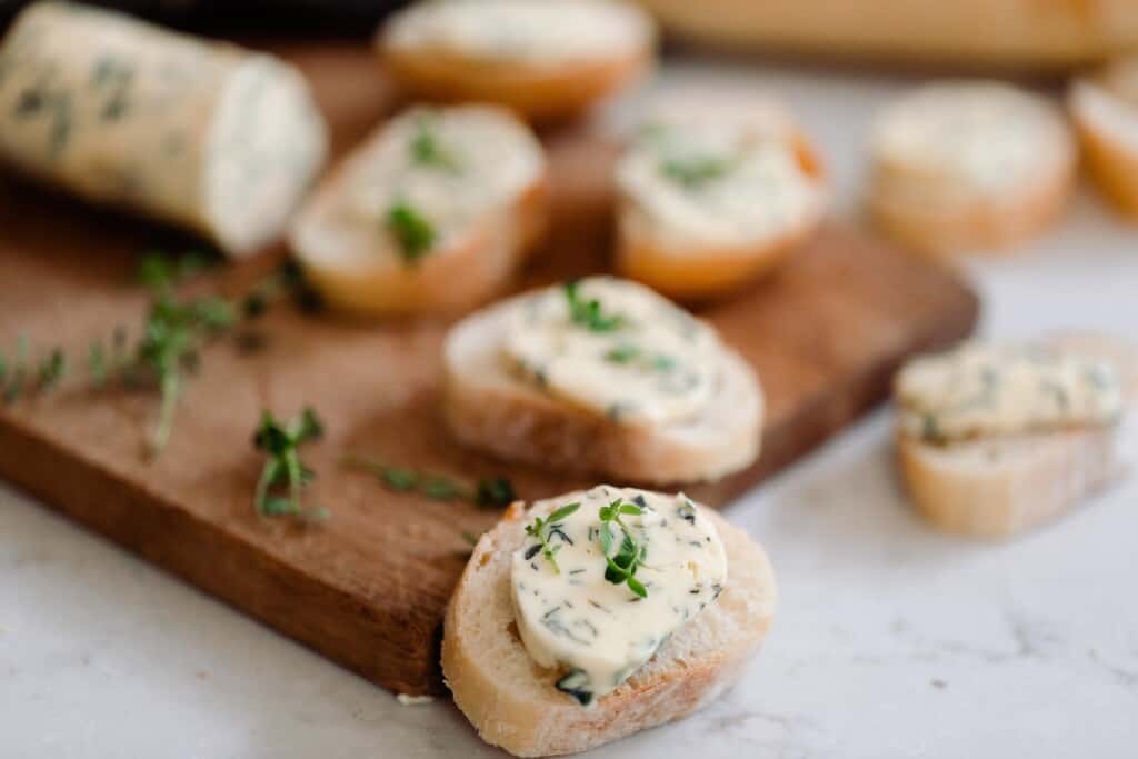 Small slices of sourdough bread with delicious herb butter on top on a brown cutting board and white marble countertop 