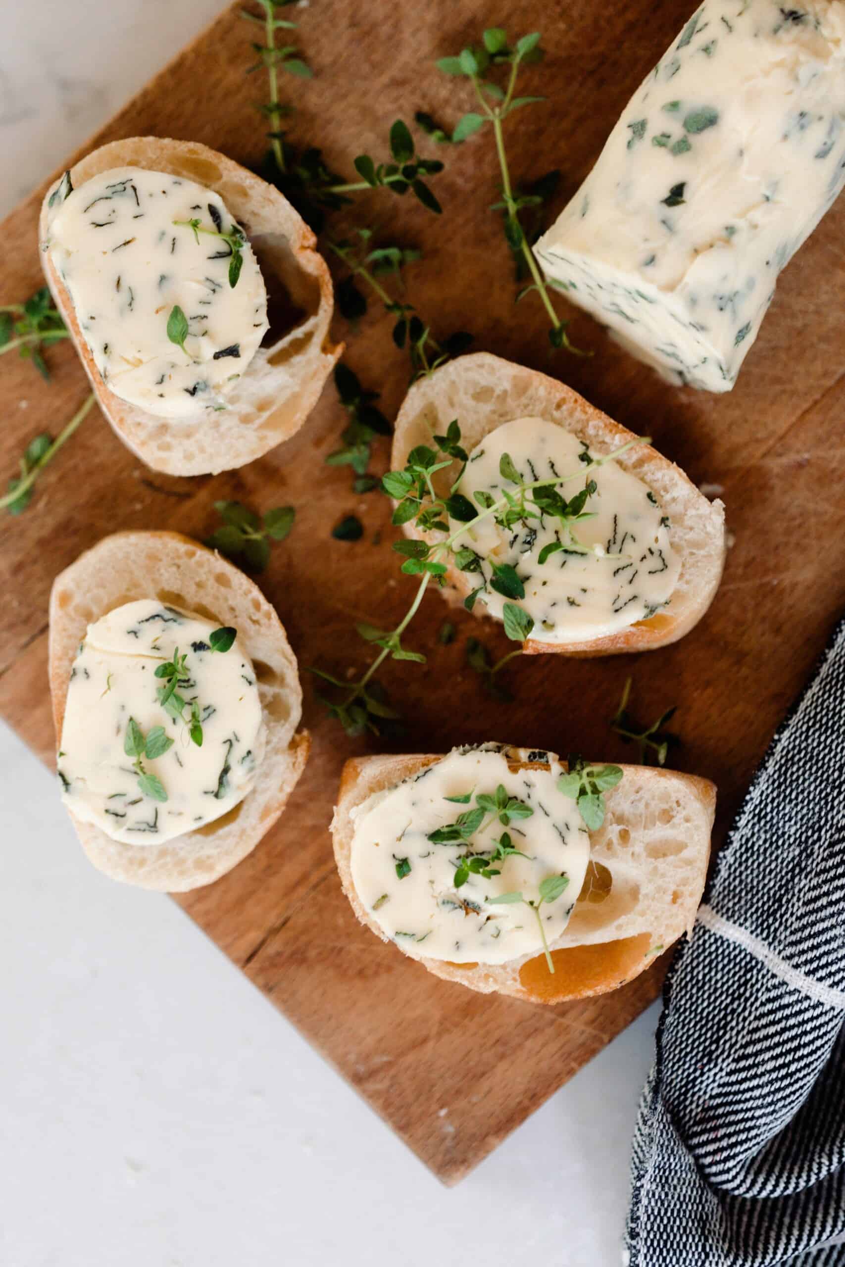 four slices of fresh bread with compound butter on top on a cutting board with herbs spread around and a log of herb butter to the back right