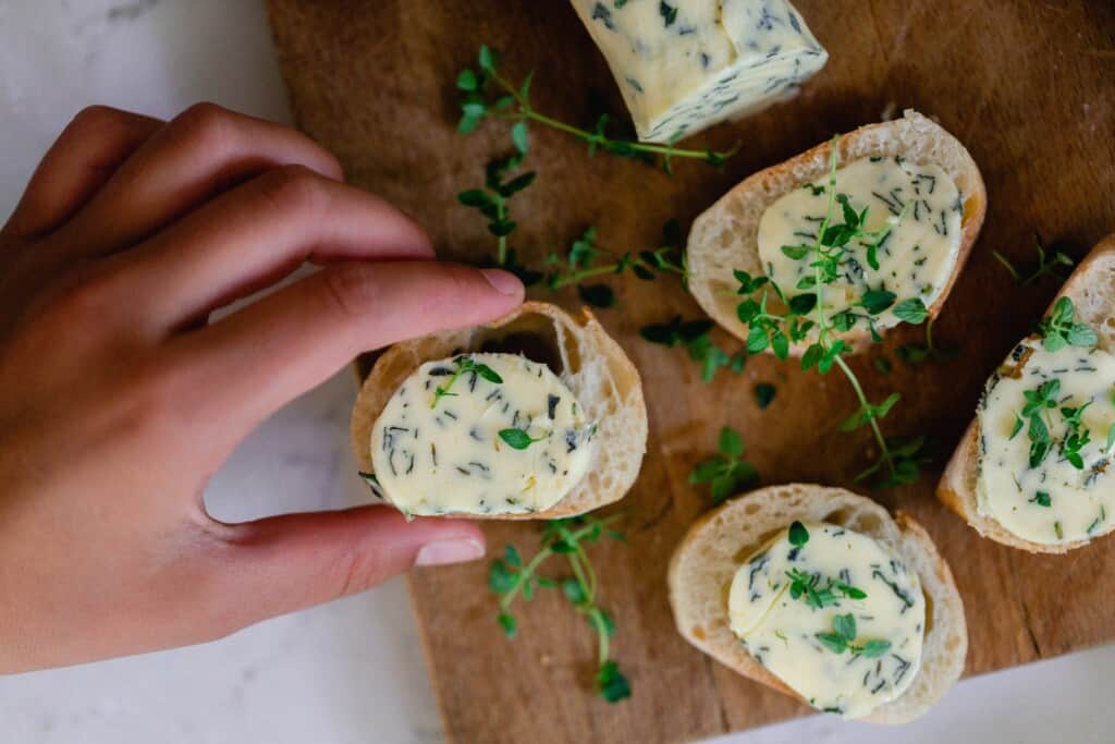 hand taking a slice of French bread topped with herb butter on a cutting board filled with more slices of toast and fresh herbs