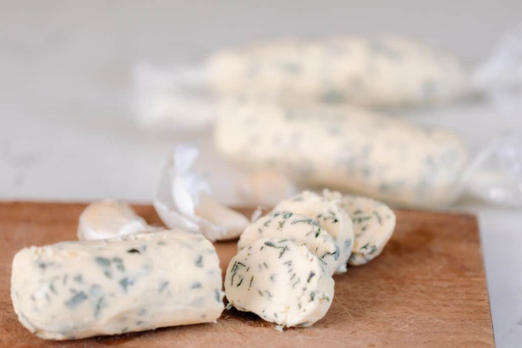 compound butter sliced on a wooden cutting board with more logs of butter wrapped in parchment paper in the background