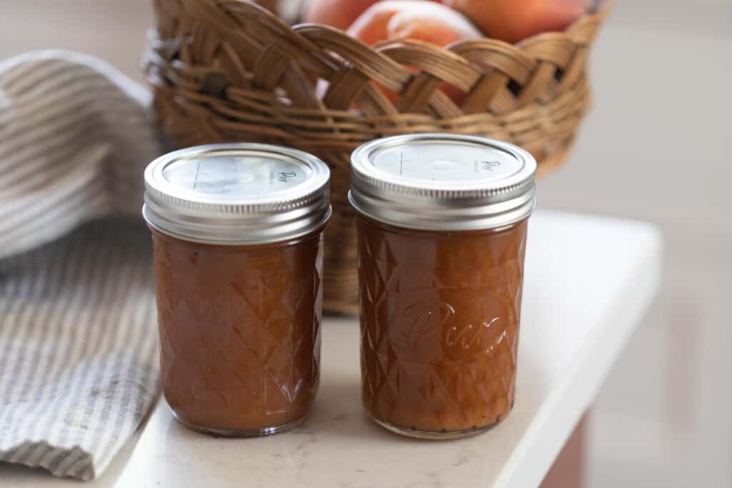 Two jars of peach preserves on a white countertop with a bake of peaches in the background.