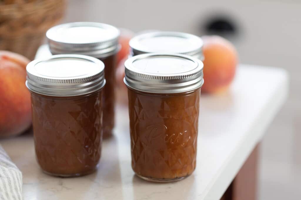 four jars of peach jam on a white countertop with peaches in the background