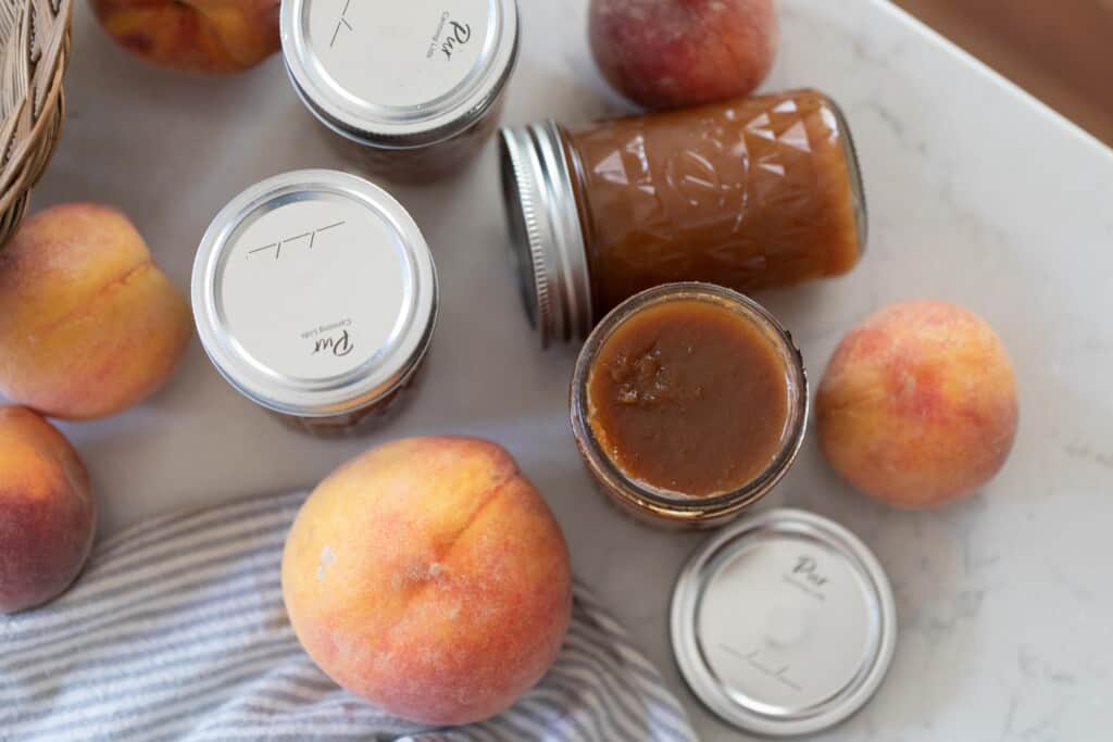 four jars of peach preserves spread out on a white countertop surrounded by peaches