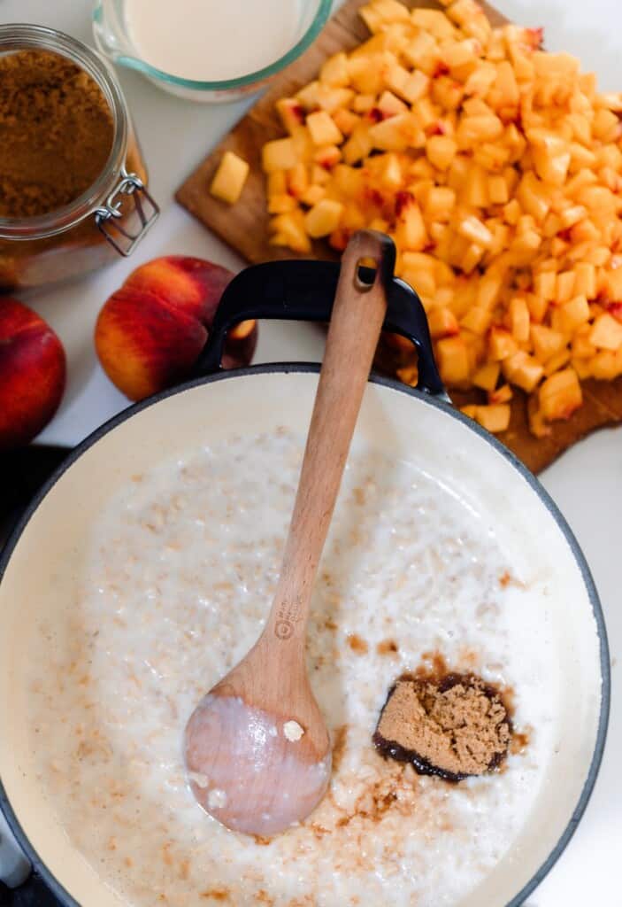 a dutch oven with milk, oats, and brown sugar on a stove with a cutting board full of chopped peaches in the background