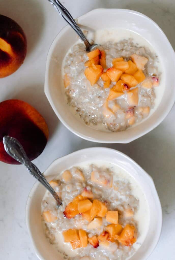 two bowls of peaches and cream oatmeal topped with extra chopped peaches on a white countertop