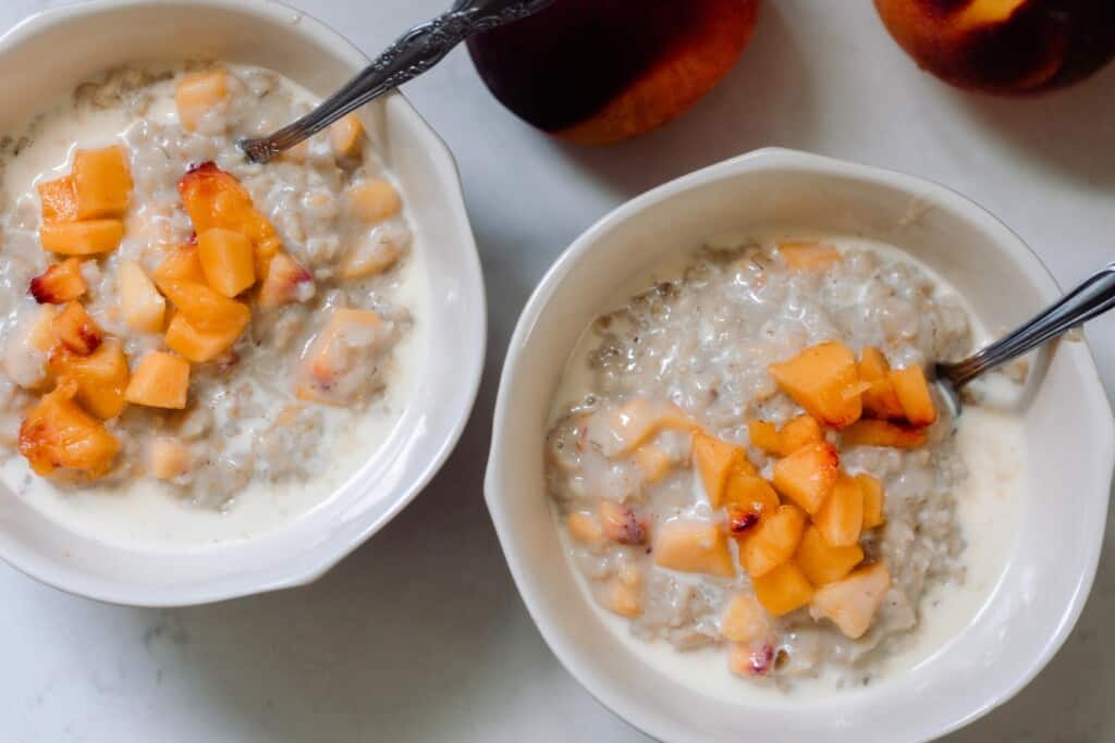 two bowls of peaches and cream oatmeal topped with peaches on a white countertop with two peaches in the top corner