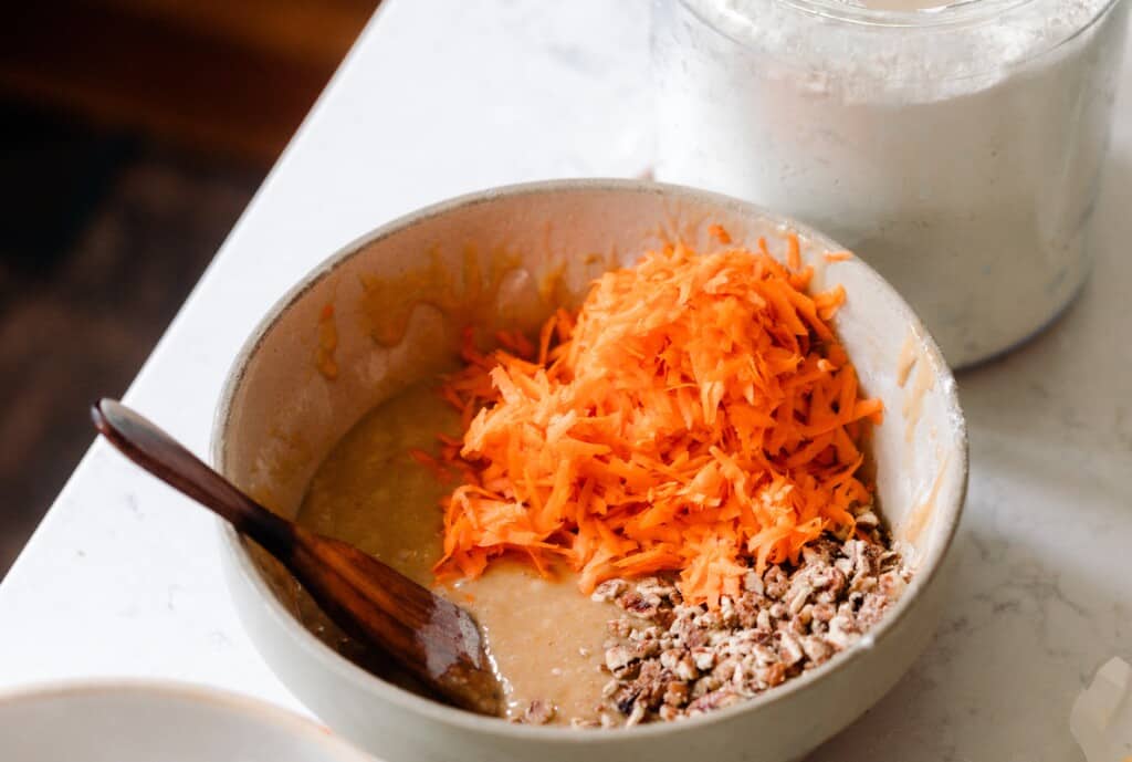 shredded carrots and chopped pecans being added to sourdough carrot cake batter
