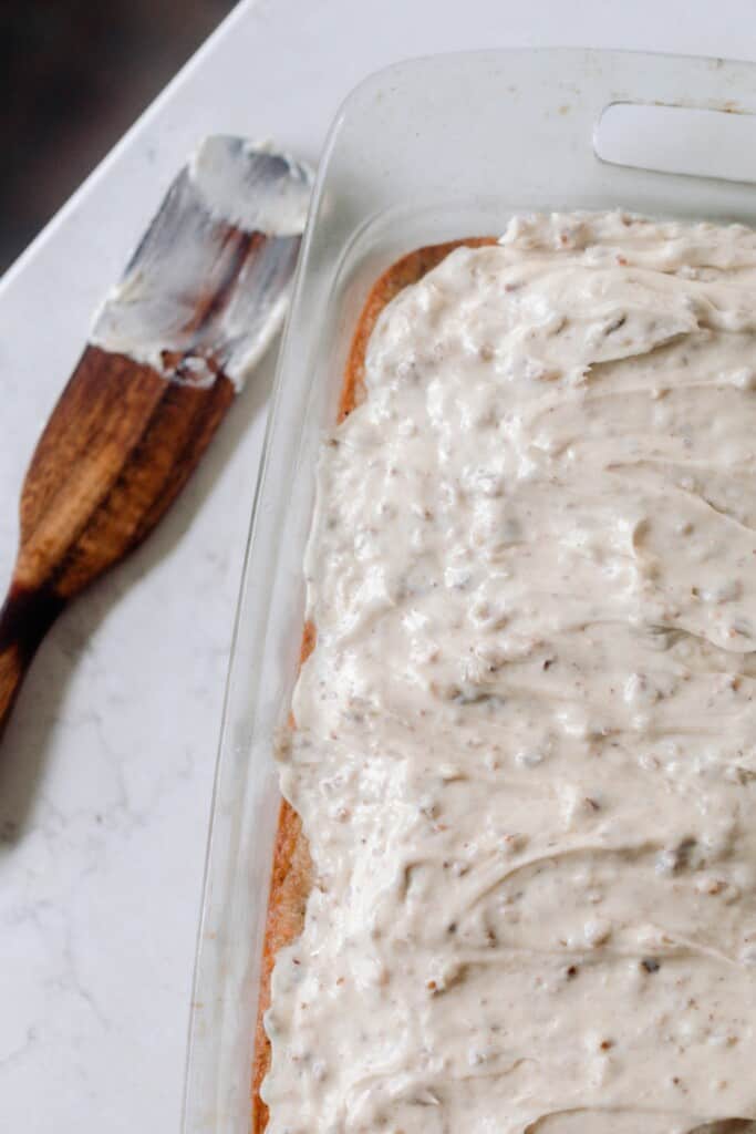 overhead photo of a sourdough carrot cake topped with cream cheese frosting in a a glass baking dish on a white countertop with a wooden spoon to the left