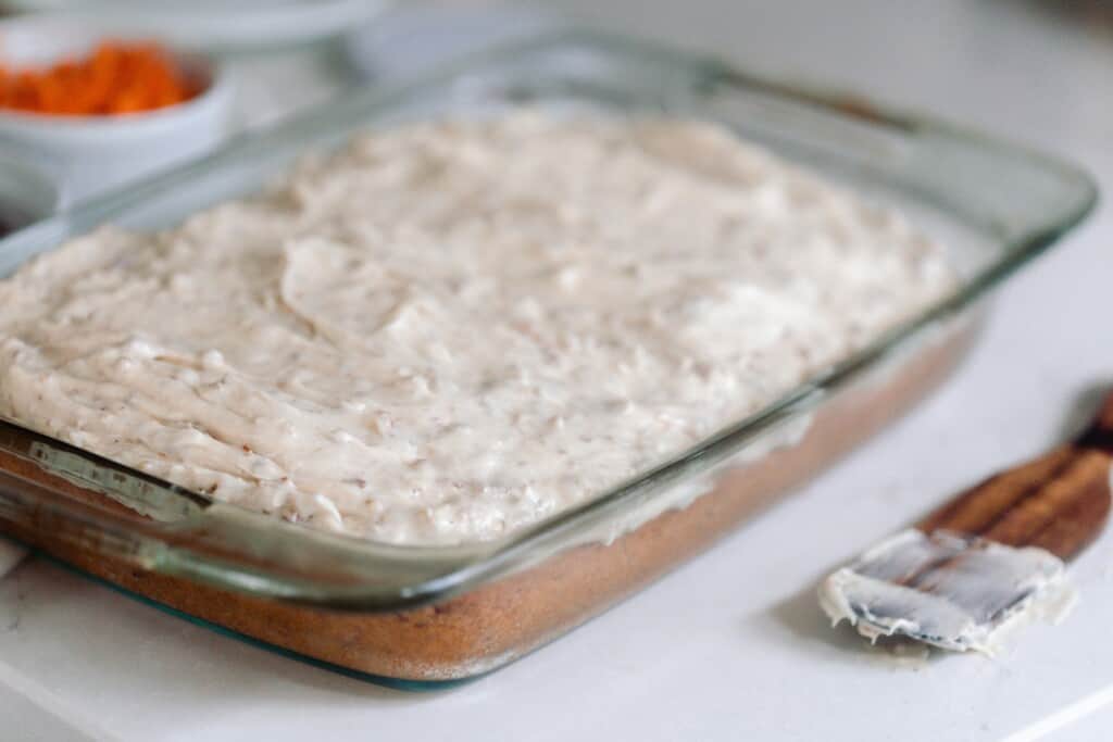 side view of a sourdough carrot cake topped with cream cheese frosting gin a glass baking dish with a wooden utensil to the right