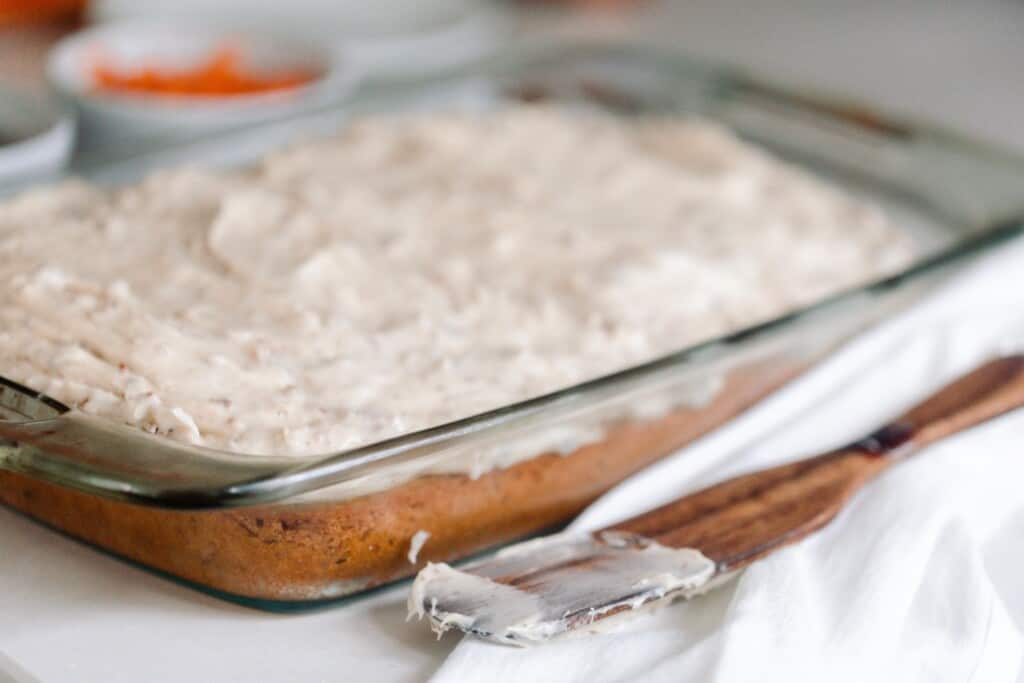 side view of sourdough carrot cake with cream cheese frosting in a glass baking dish with a wooden utensil covered with frosting to the right