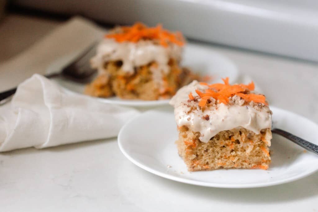 two slices of sourdough carrot cake topped with cream cheese frosting and a few shredded carrots and chopped pecans on white plates on a white countertop