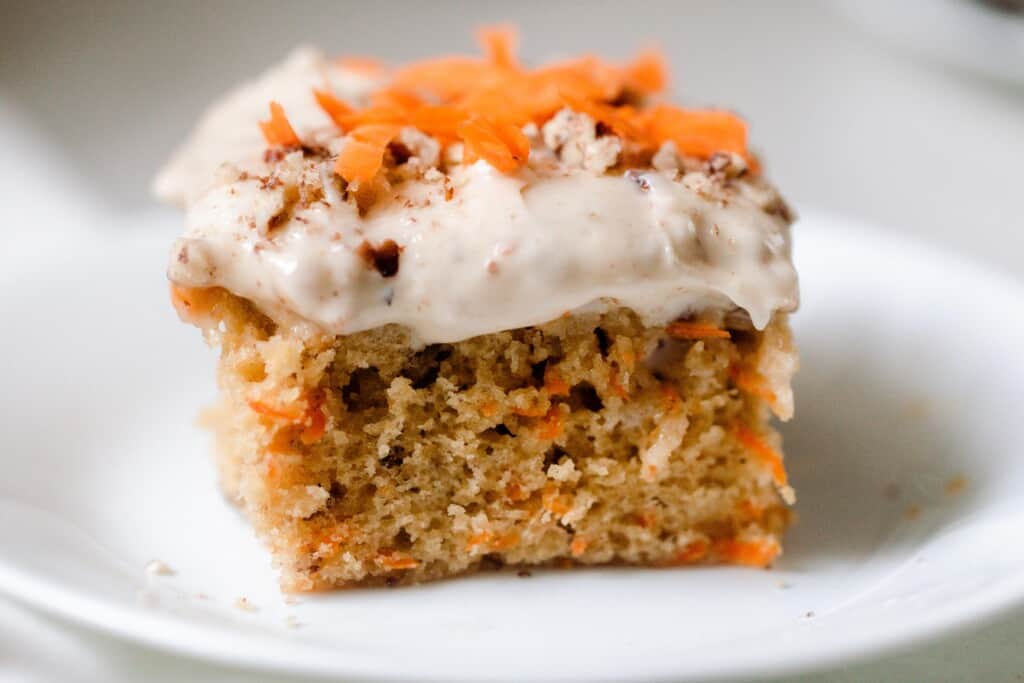 close up picture of a square slice of sourdough carrot cake topped with cream cheese frosting, shredded carrots and pecans