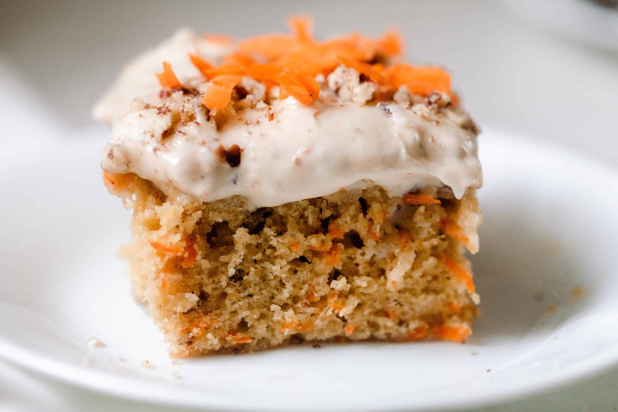 close up picture of a square slice of sourdough carrot cake topped with cream cheese frosting, shredded carrots and pecans