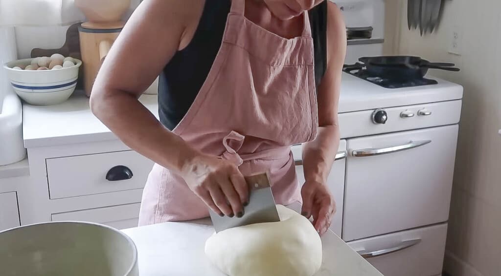 woman wearing a pink apron using a bench scraper to cut a ball of dough in half