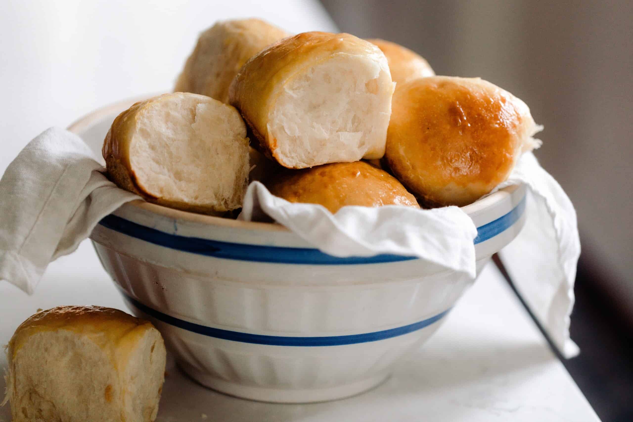 sourdough Hawaiian sweet rolls stacked in a white and cream ironstone bowl lined with a white linen towel. The bowl is on a white countertop