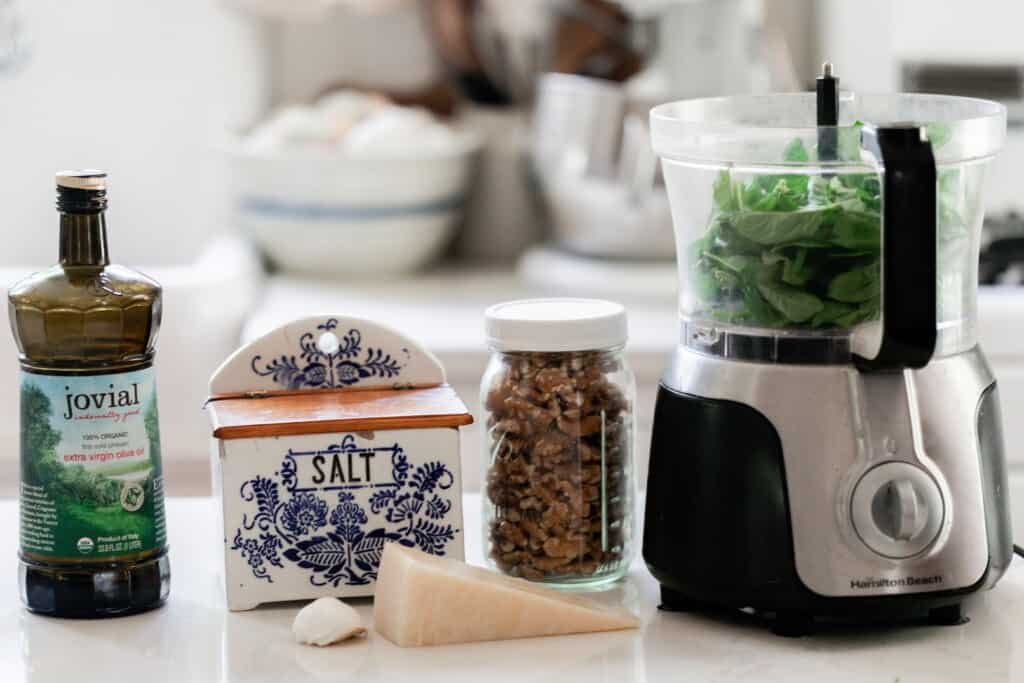 olive oil, salt, Parmesan, garlic, walnuts in a jar, and a food processor with basil on a white countertop in a kitchen