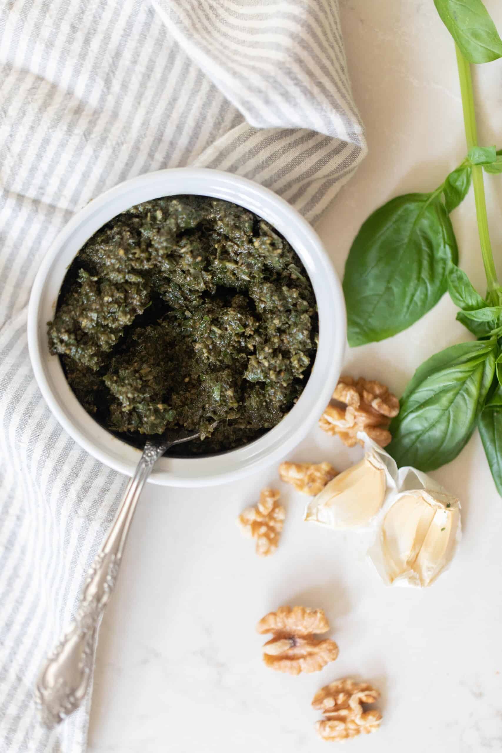 overhead photo of walnut pesto in a white jar with a spoon on a white countertop with walnuts and basil spread around the counter.