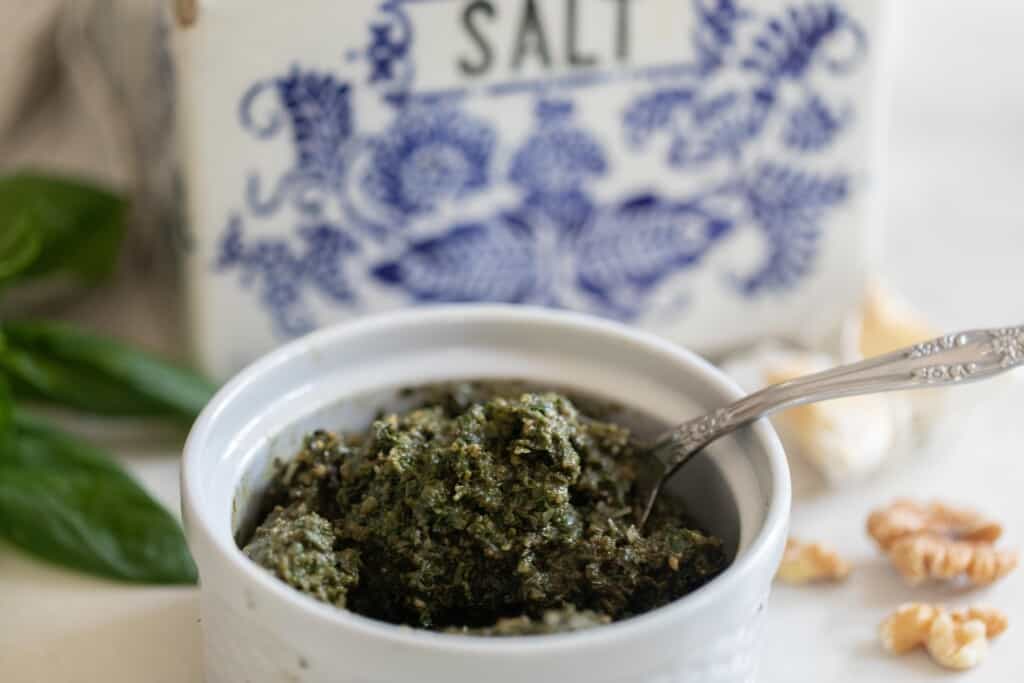 close up side view of a white ceramic jar with pesto and a spoon in the jar. A white and blue vintage salt box is in the background.