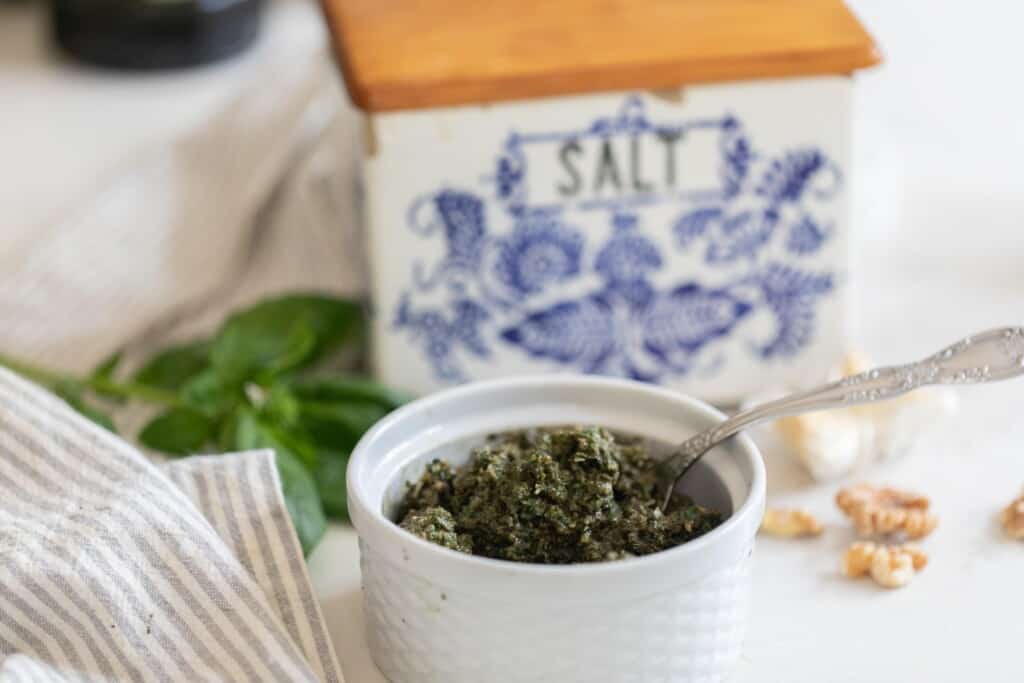 a white ramekin filled with pesto on a white counter with walnuts and basil spread about the countertop and a vintage box of salt