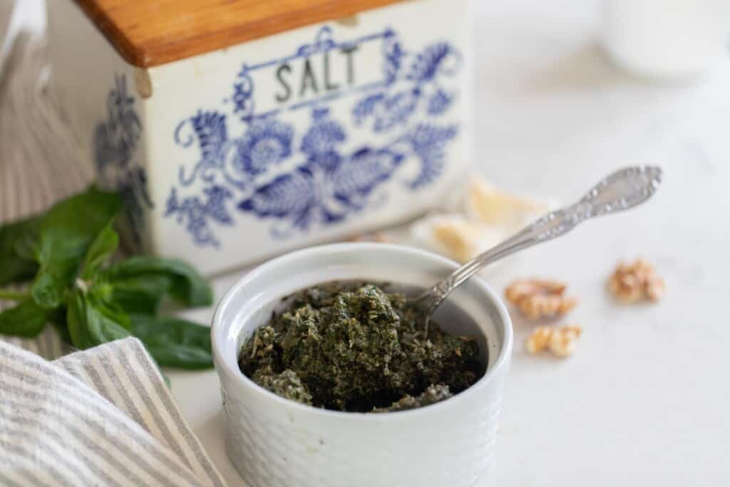 pesto in a white ramekin with a spoon on a white countertop with walnuts basil, a stripped towel and a vintage blue and white salt box in the background