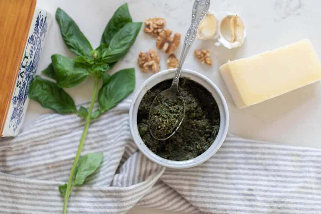 overhead photo of walnut pesto in a jar with a spoon on a white countertop surrounded bye basil, walnuts, garlic cloves, Parmesan cheese and a stripped tea towel.