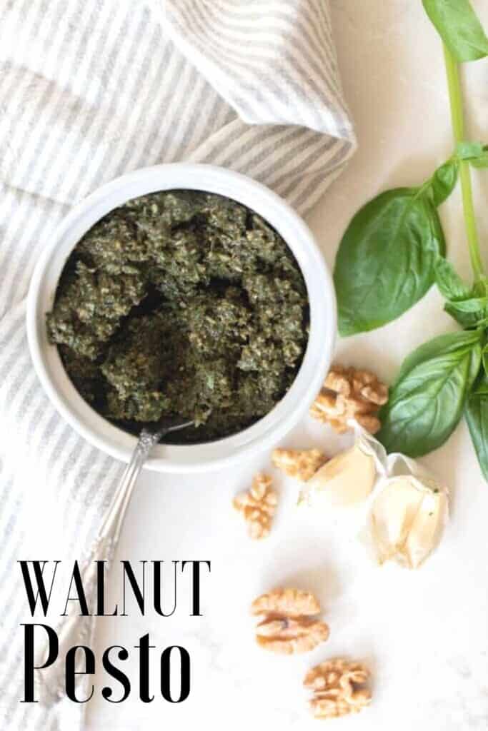 overhead photo of walnut pesto in a white jar with a spoon on a white countertop with walnuts and basil spread around the counter.