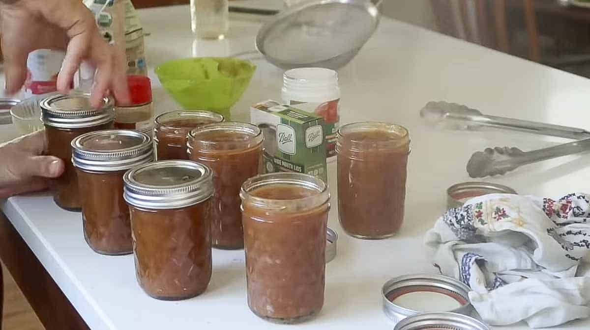 Woman tightening lid and ring onto a jar of peach preserves with more jars and tools around the island.