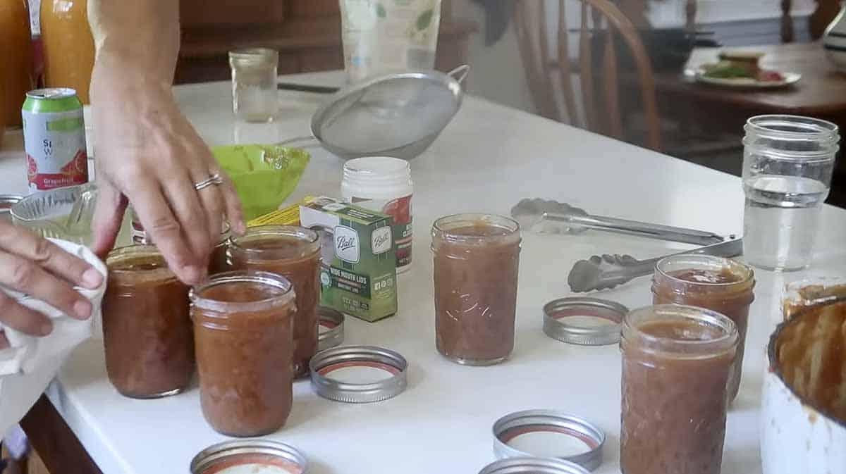 Woman wiping the lid of a jar of peach preserves with a clean rag, on a table of canning jars and supplies.