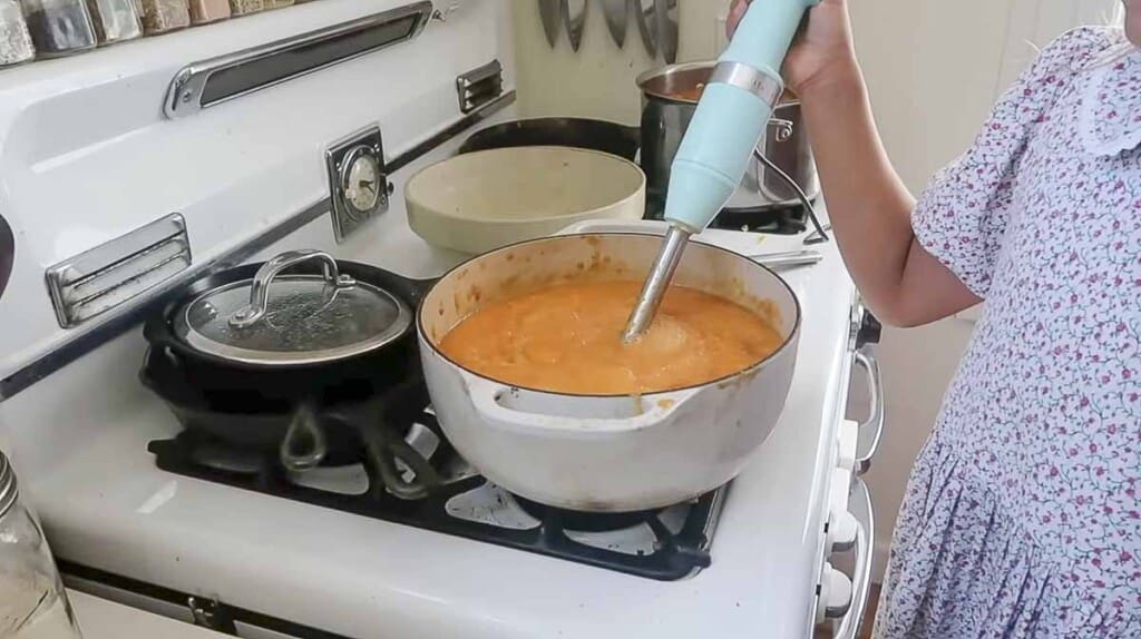 peaches being blended with a immersion blender in a white dutch oven on a white antique stove