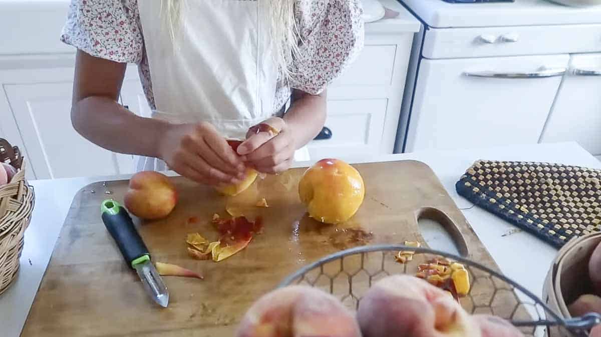 Girl in an apron peeling peaches on a wood cutting board.