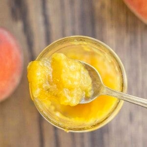 overhead photo of a spoonful of peach preserves on top of a jar of preserves. The jar rests on a wood table surrounded by peaches