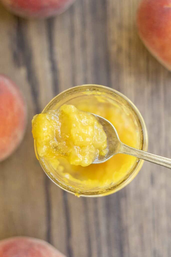 overhead photo of a spoonful of peach preserves on top of a jar of preserves. The jar rests on a wood table surrounded by peaches