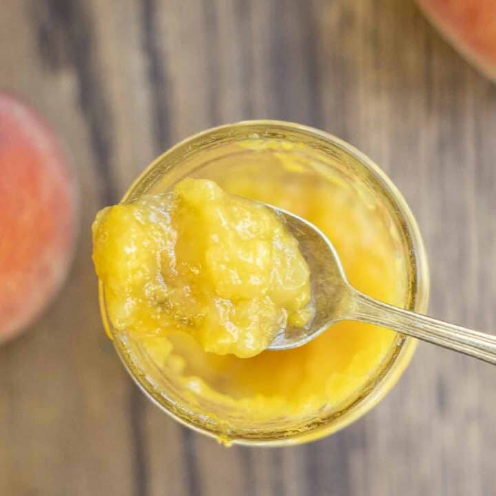 overhead photo of a spoonful of peach preserves on top of a jar of preserves. The jar rests on a wood table surrounded by peaches