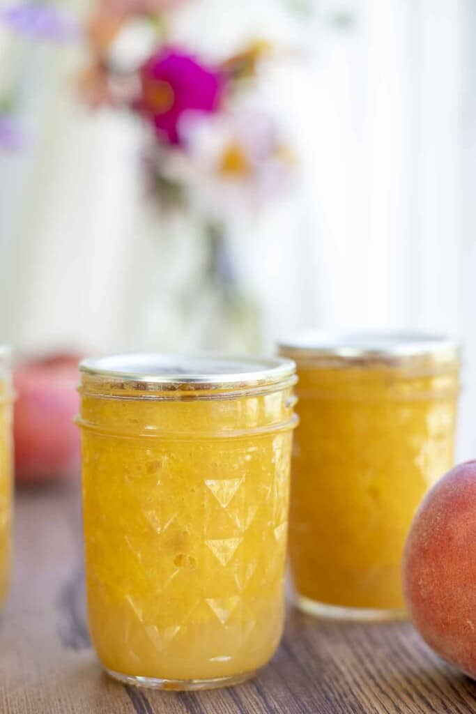two jars of peach jam on a wood table with peaches around the jar and a vase of flowers in the background