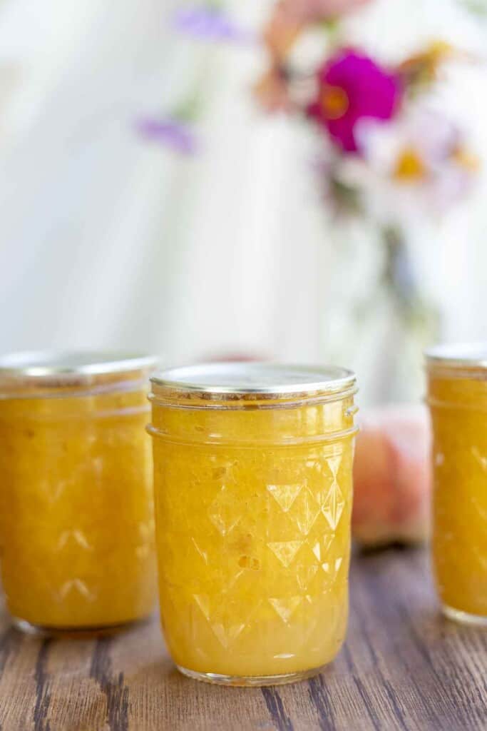 three jars of peach jam on a wood table with a vase of colorful flowers in the background