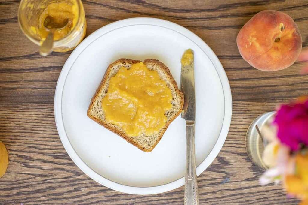 slice of toast with peach preserves with a jam filled knife on a white plate on a wood table. A jar of jam, a fresh peach, and a vase of flowers surrounding the plate