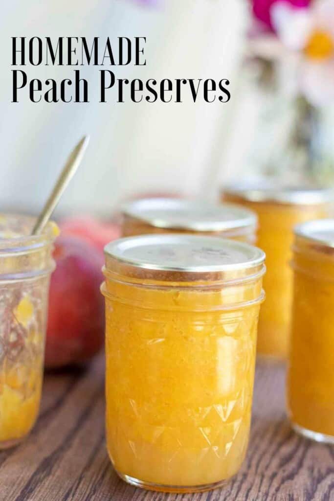 jars of canned peach preserves, including an opened jar with a knife, on a wood table. Peaches and flowers are in the background