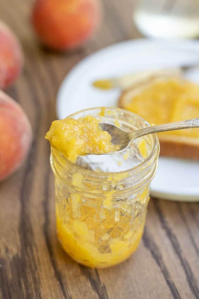 spoonful of jam resting on a jar of peach jam with a plate with toast covered jam and fresh peaches behind the jar