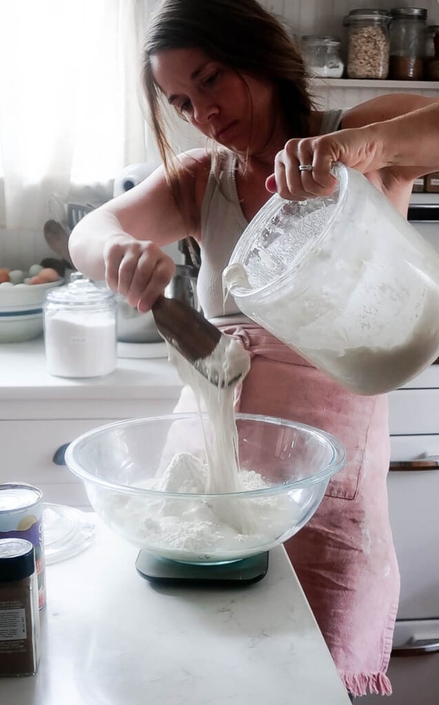 women wearing a red apron adding sourdough starter to a glass bowl on a kitchen scale on a white countertop