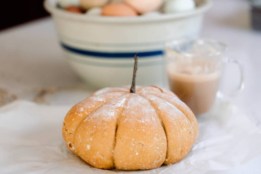 pumpkin shaped sourdough bread on a white quartz countertop with a glass mug of coffee and a large bowl of eggs in the background