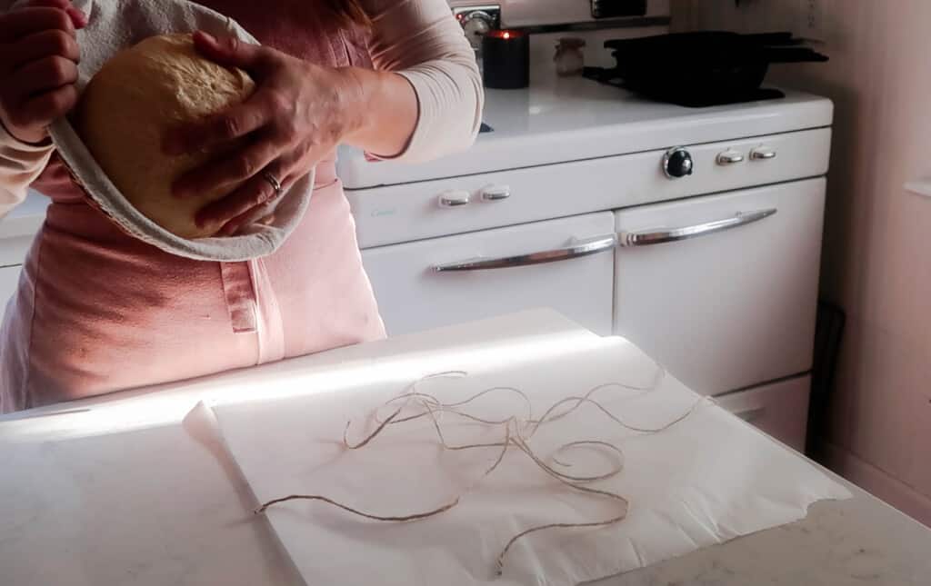 a woman placing a sourdough boule onto a piece of parchment paper with string lined up on the parchment