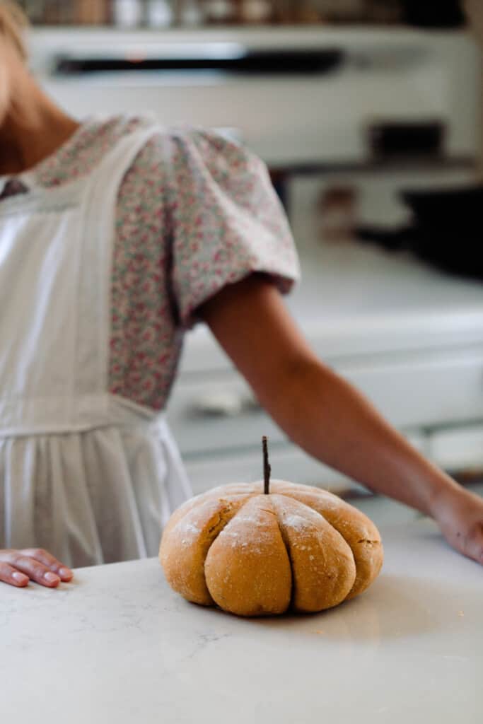 girl standing next to a pumpkin shaped loaf of sourdough bread laying on a white quartz counter. A vintage stove in is the background