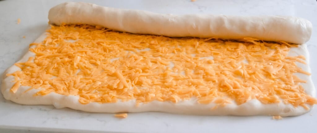 bread dough rolled into a rectangle, topped with shredded cheese and is being rolled up.