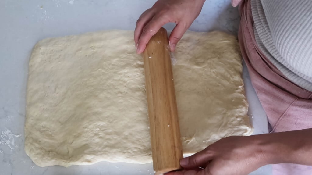 bread dough being rolled out into a rectangle with a rolling pin on a white countertop