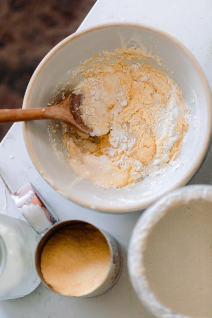 a bowl of cornmeal, flour, sourdough starter, sugar, and milk with measuring cups around