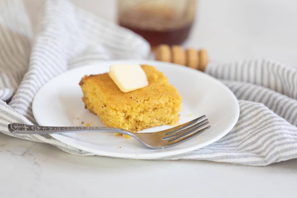 square slice of sourdough cornbread with a pat of butter on top on a white plate with a fork resting on the plate. The plate sits on a white and blue stripped towel on a white countertop